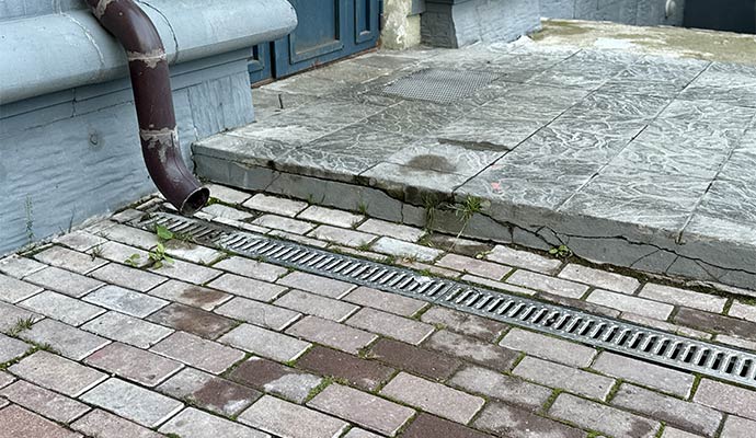 A dark brown gutter downspout emptying water into a metal grate trench drain